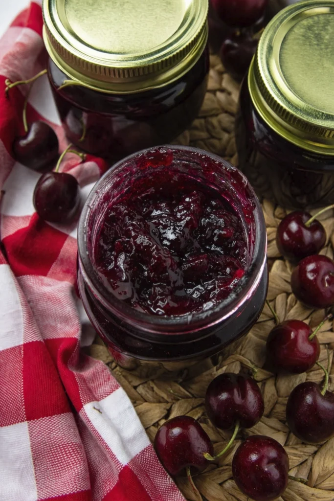 Homemade cherry jam in a mason jar with fresh cherries, gingham towel, and rustic kitchen styling
