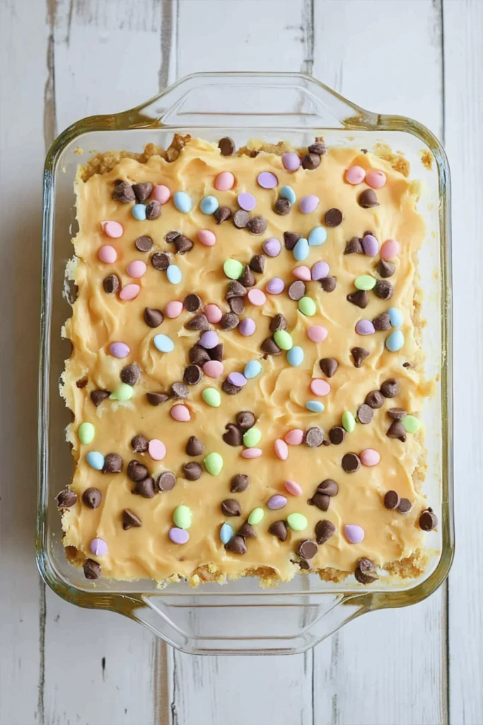 Pastel Easter dessert bars with orange frosting, mini chocolate chips, and colorful candy sprinkles in a glass baking dish.