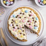 Close-up of a thick chocolate chip cookie cake slice with melted chocolate chunks and a peanut butter cup center, topped with piped vanilla buttercream and pastel candy sprinkles.
