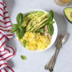 Healthy breakfast bowl with scrambled eggs, sliced avocado, grains, basil garnish, and chili flakes on a light background
