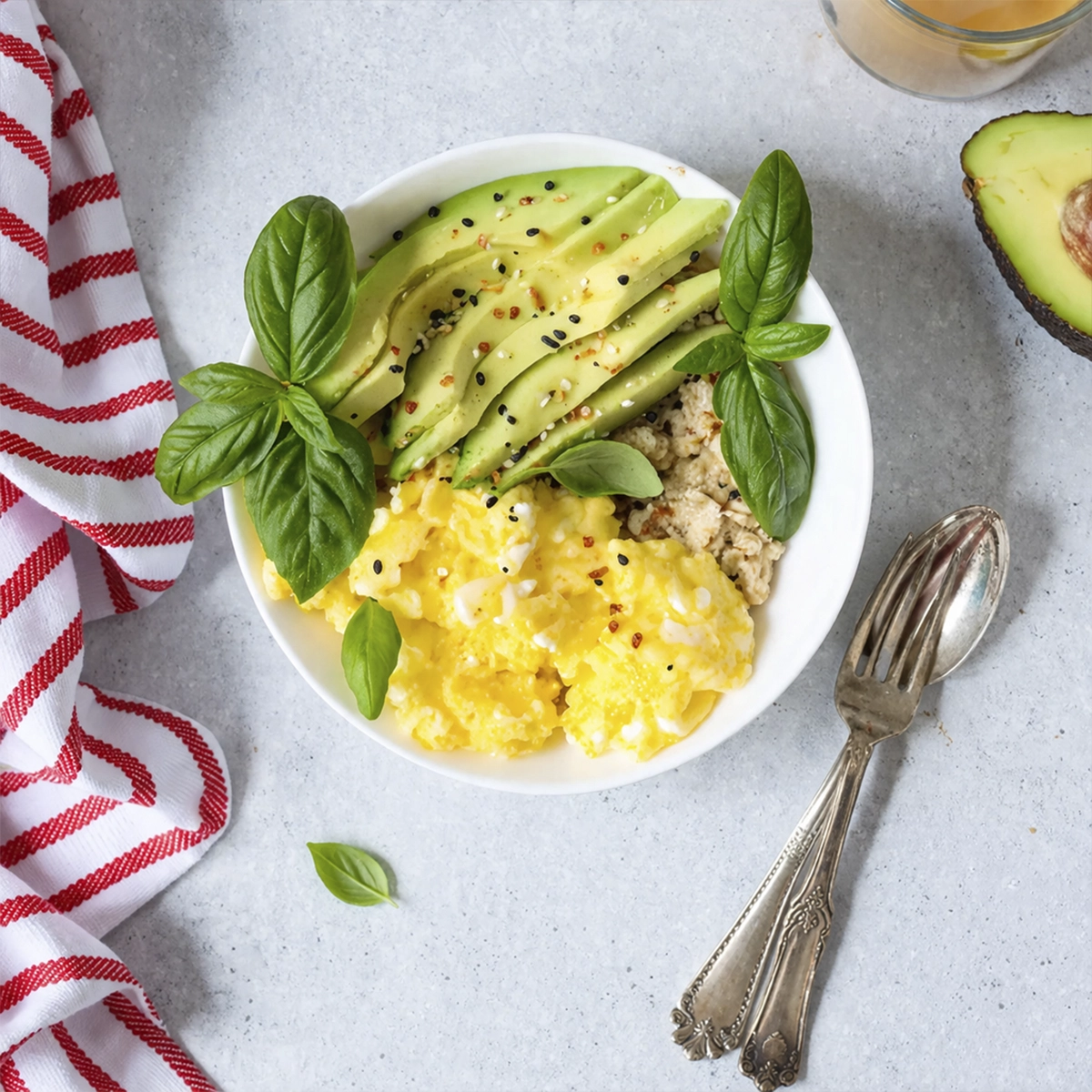 Healthy breakfast bowl with scrambled eggs, sliced avocado, grains, basil garnish, and chili flakes on a light background