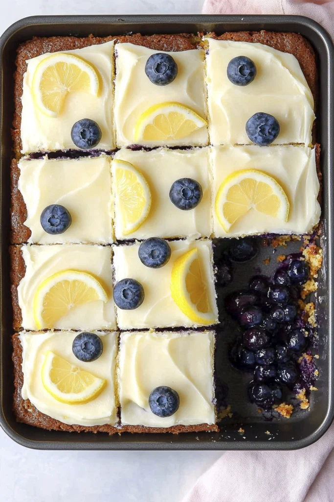 Lemon blueberry sheet cake bars with cream cheese frosting topped with lemon slices and fresh blueberries in a baking pan.