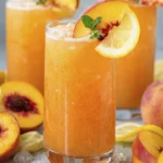 Tall glass of peach lemonade with ice, peach slice, lemon wheel, and mint garnish surrounded by fresh peaches and lemon wedges.