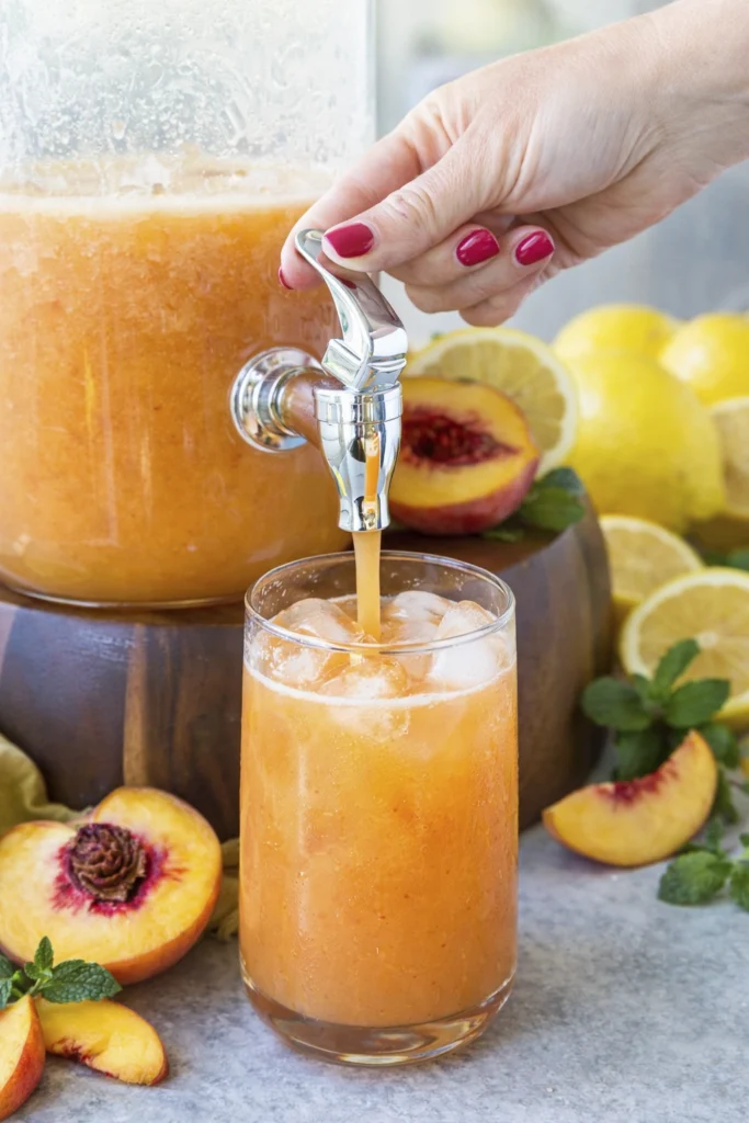 Hand pouring peach lemonade mocktail from a glass dispenser into an iced glass surrounded by peaches, lemons, and mint.
