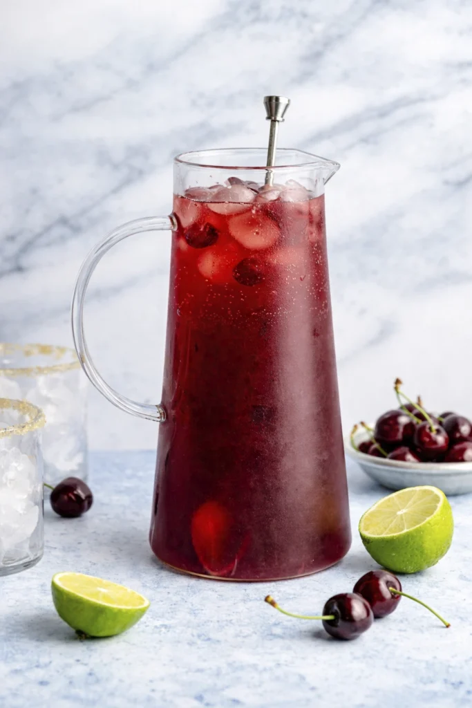 Tall glass pitcher filled with cherry lime mocktail, ice, fresh cherries, and lime halves styled on a light blue surface with a marble background.