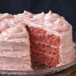 Slice of strawberry layer cake with pink strawberry frosting on a white plate with the whole cake in the background.