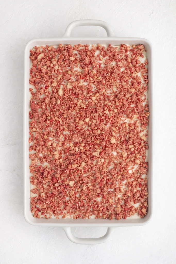 Top-down view of a baking dish with strawberry crumble topping spread over a creamy dessert base.