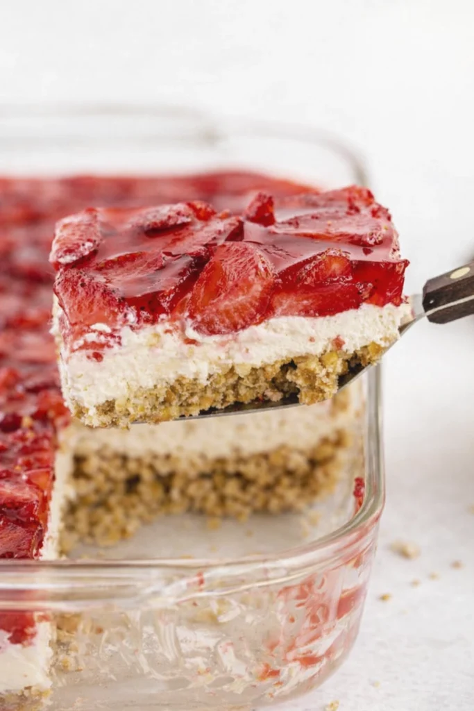 Close-up of a strawberry cheesecake bar with gelatin topping being lifted from a glass dish, showing creamy and crumb layers.