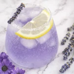 Lavender lemonade in a glass with lemon slice, sugared rim, ice cubes, and fresh lavender garnish on a marble surface.