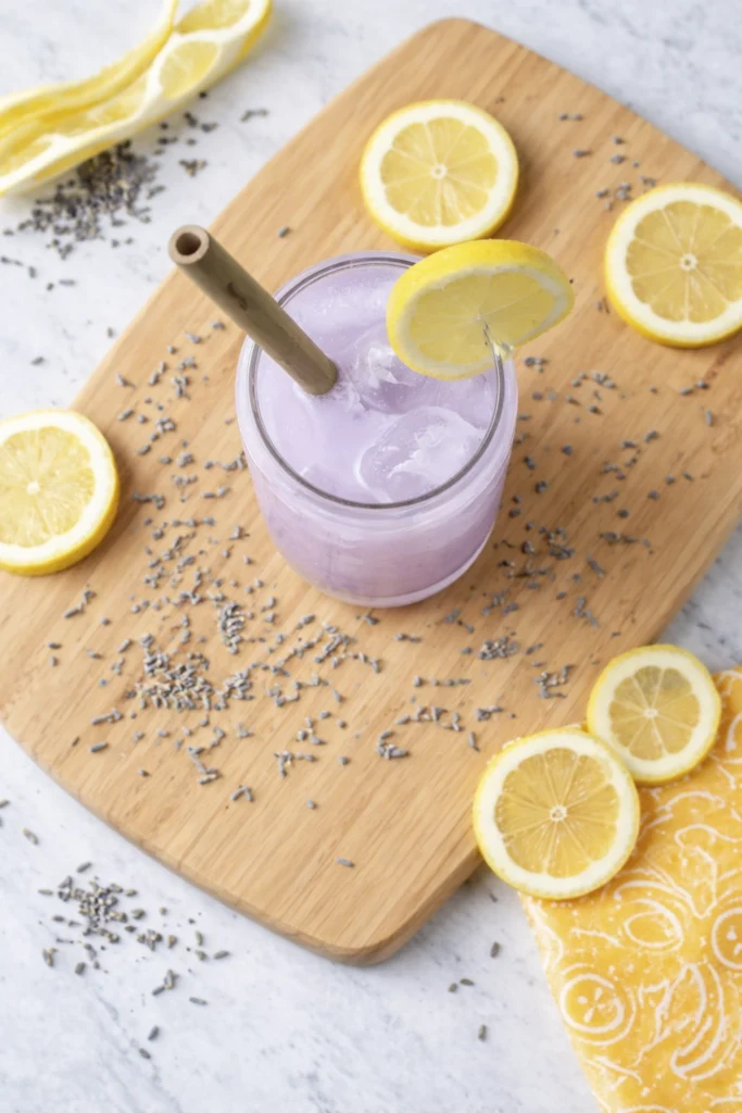 Top-down view of a pastel lavender lemonade mocktail with ice, lemon garnish, straw, dried lavender, and lemon slices on a wooden board.