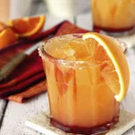 Fresh orange mocktail in a short glass with ice, sugar rim, and orange slice on a wooden coaster.