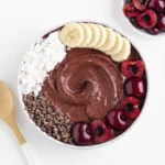 Chocolate cherry smoothie bowl topped with banana slices, fresh cherries, coconut flakes, and cacao nibs on a white background.
