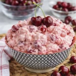 Bowl of creamy pink cherry fluff salad topped with fresh cherries and surrounded by whole cherries on a rustic table setting.
