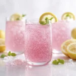Sparkling pink lemonade in clear glasses with lemon slices and mint garnish, styled with fresh lemons on a bright white background.