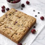 Cherry chocolate chip blondie bars cut into squares on parchment paper with fresh cherries and a white bowl of cherries on a marble background.