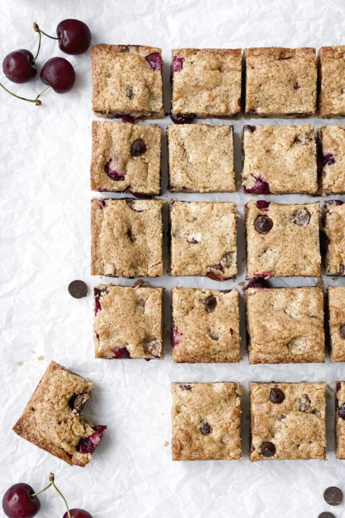 Overhead photo of cherry chocolate chip blondie bars cut into squares on white parchment paper with whole cherries and one bitten blondie piece.