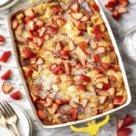 Overhead view of strawberry French toast casserole baked in a ceramic dish, topped with fresh strawberries and powdered sugar on a marble surface.