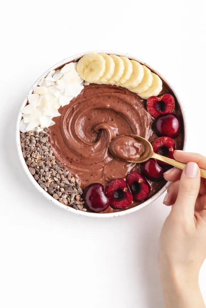 Top-down view of a chocolate cherry smoothie bowl with banana slices, coconut flakes, cacao nibs, fresh cherries, and a gold spoon taking a scoop.