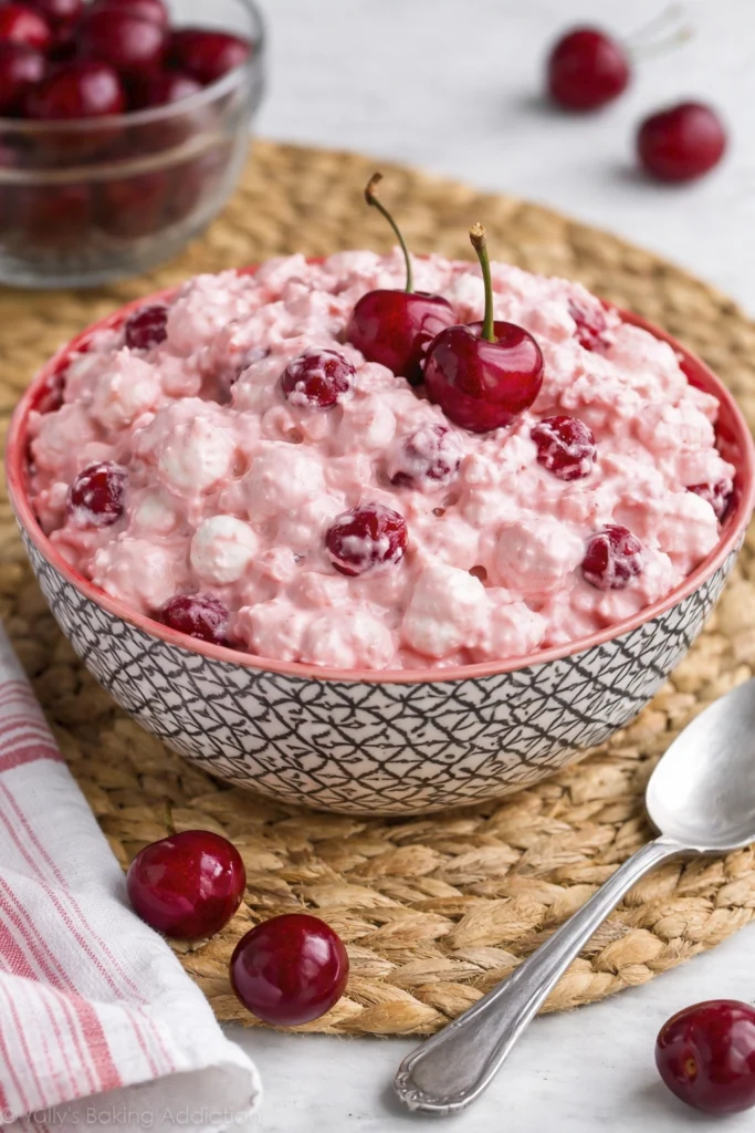 Bowl of creamy pink cherry fluff salad topped with fresh cherries and surrounded by whole cherries on a woven placemat.