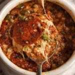 Close-up of a spoon lifting cheesy baked casserole with tomato sauce and herbs from a white ramekin