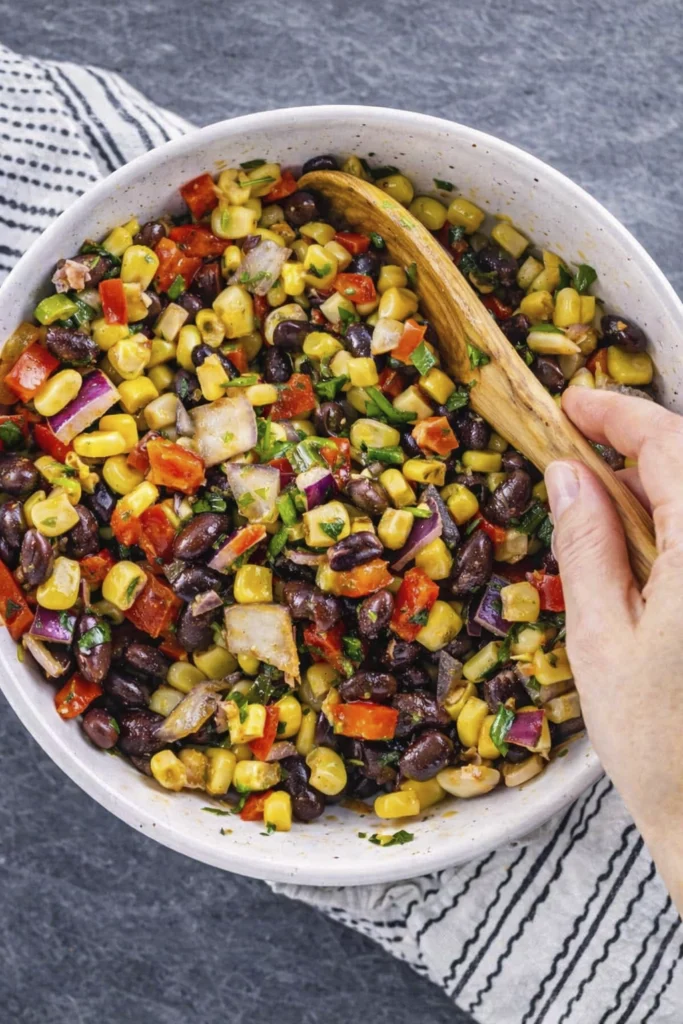 Overhead view of black bean and corn salad with red onion, bell peppers, and herbs in a white bowl with a wooden spoon.