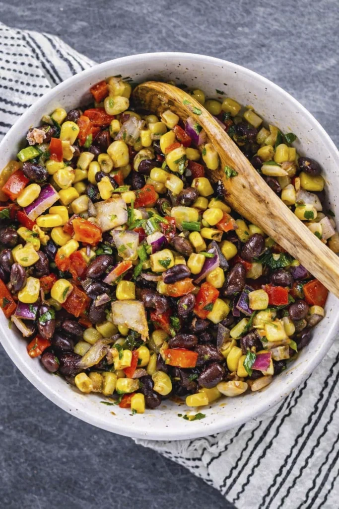 Overhead view of black bean and corn salad with red onion, bell peppers, and herbs in a white bowl with a wooden spoon.