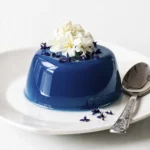 Glossy blue mirror glaze mousse dessert topped with white edible flowers and purple petals on a white plate with a spoon.