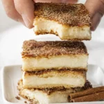 Stacked creamy custard dessert bars with cinnamon sugar topping and a bitten square held above on a white plate.