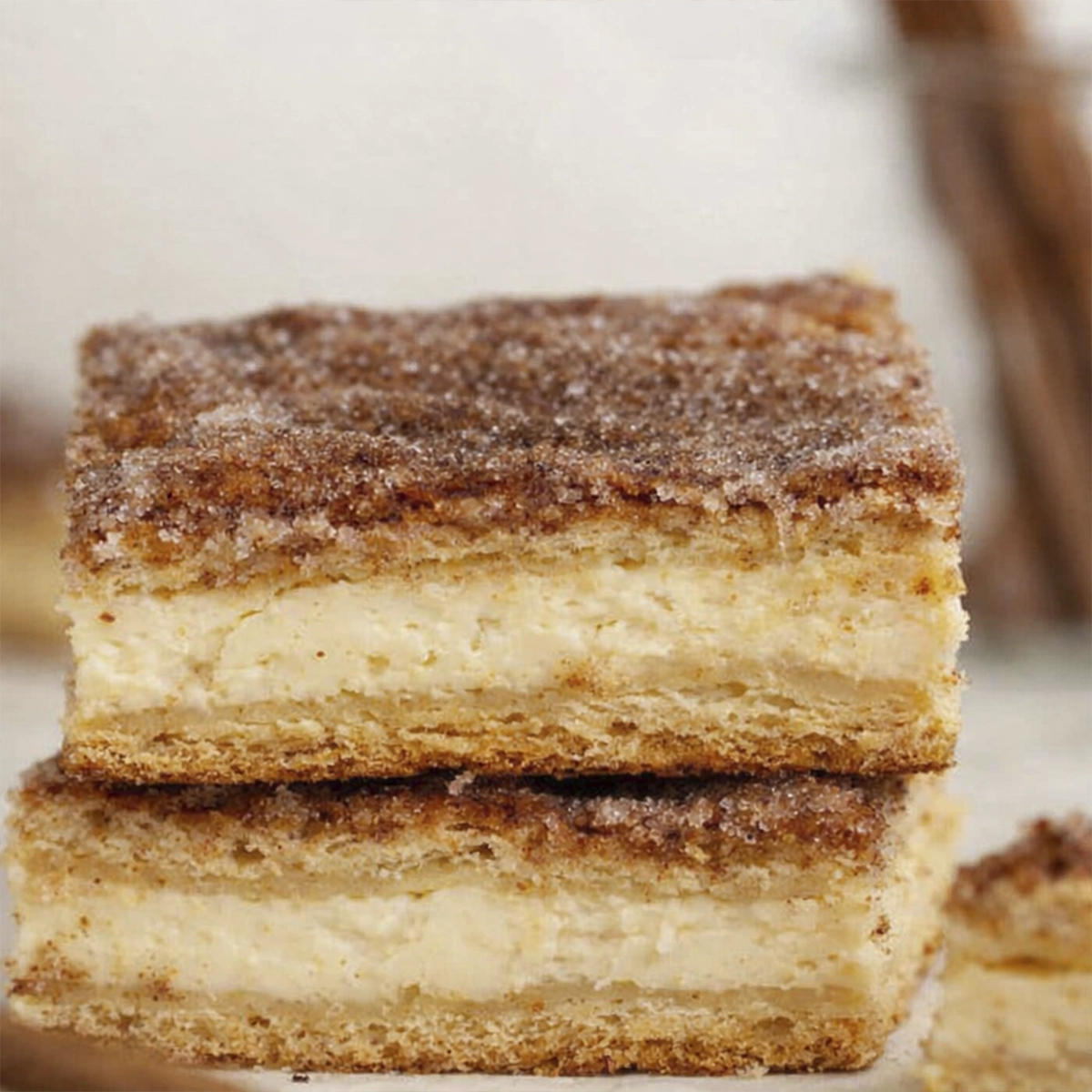 Creamy cinnamon cheesecake bars with golden layers and sugar crust stacked on a plate with cinnamon sticks in the background