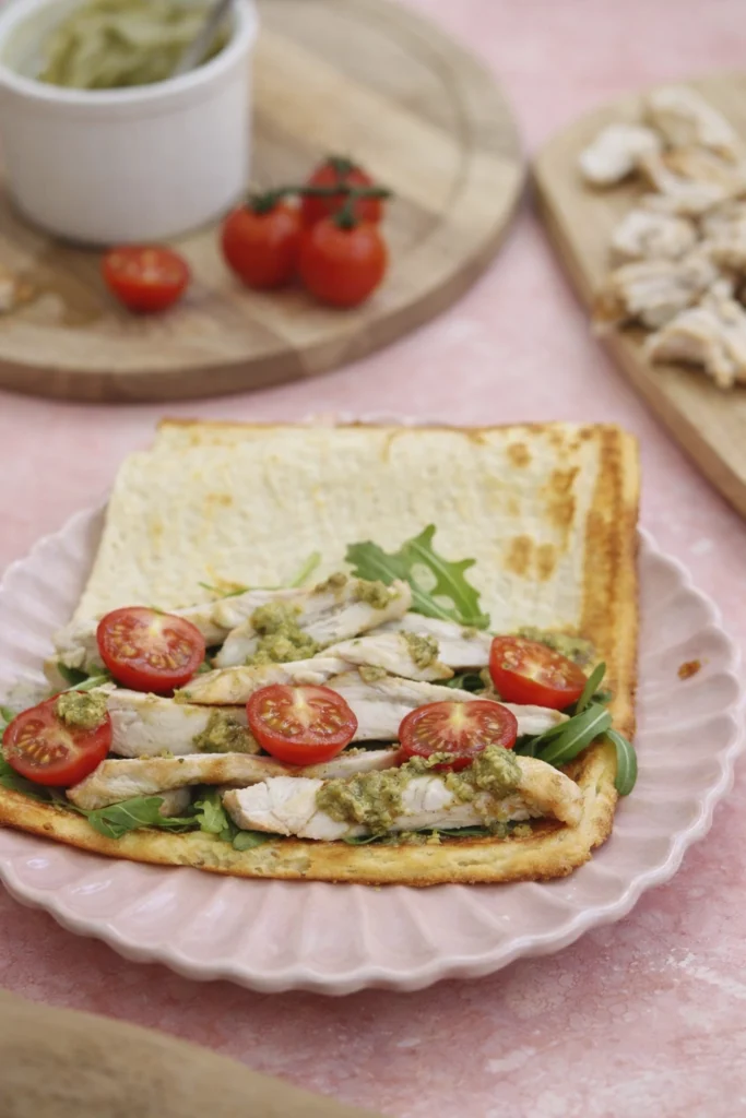 Open savory crepe with grilled chicken, cherry tomatoes, arugula, and pesto on a pink plate