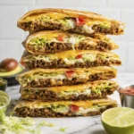 Stacked cheesy beef quesadillas with lettuce, tomatoes, and melted cheese served with lime and salsa.