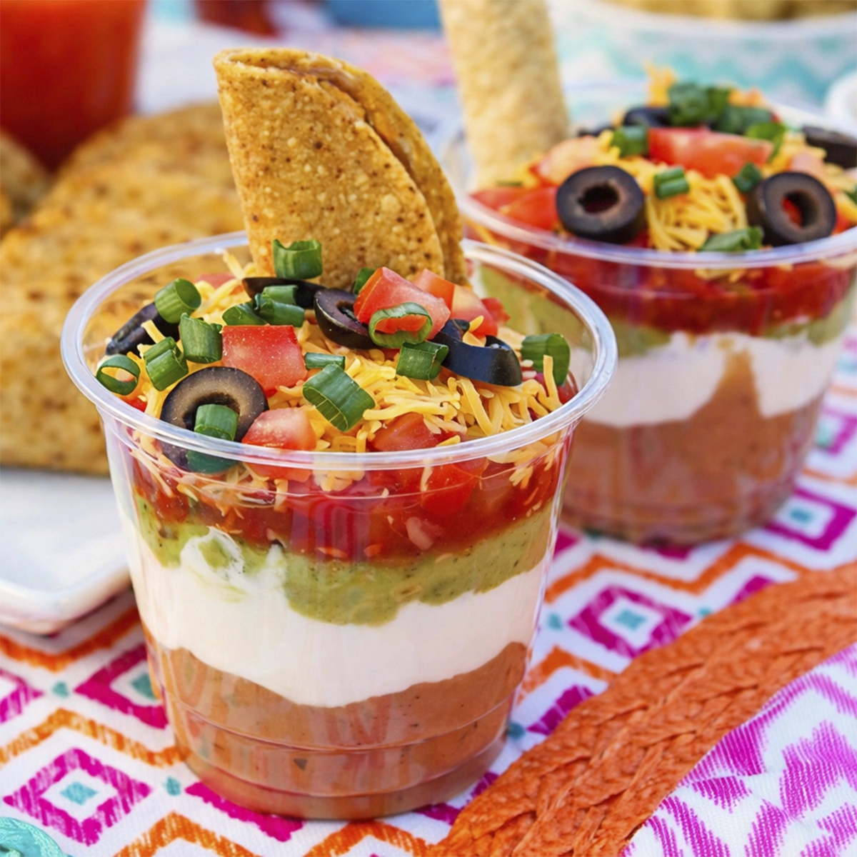 Layered taco dip cups with beans, sour cream, guacamole, salsa, cheese, olives, and tortilla chips on a colorful tablecloth