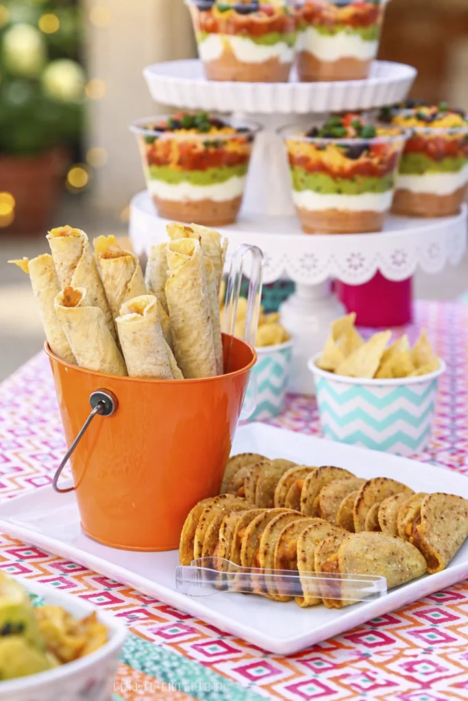 Layered taco dip cups with beans, sour cream, guacamole, salsa, cheese, olives, and tortilla chips on a colorful tablecloth