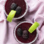 Overhead view of two blackberry frozen margaritas with sugared rims, lime wedges, and fresh blackberries on striped cloth.