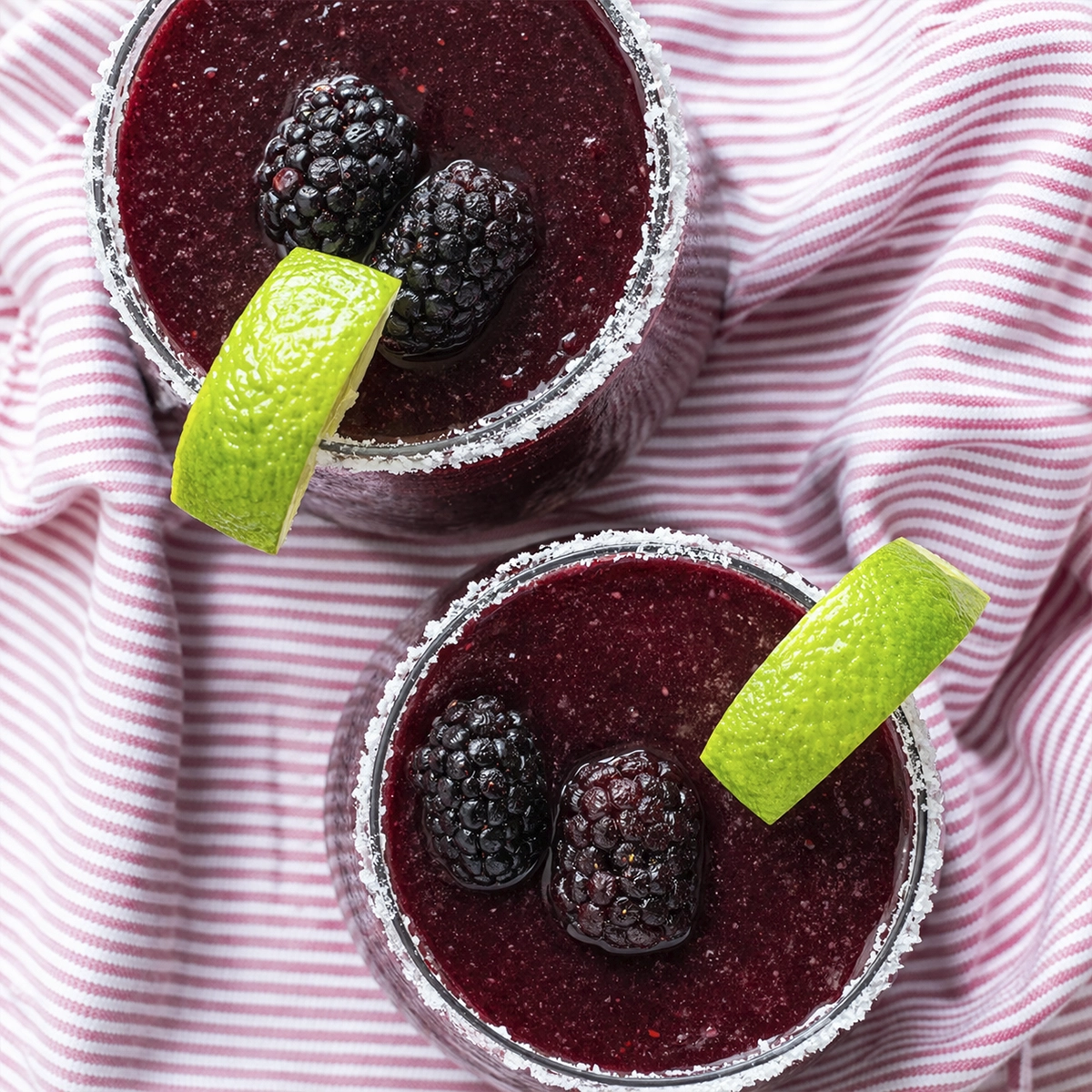 Overhead view of two blackberry frozen margaritas with sugared rims, lime wedges, and fresh blackberries on striped cloth.