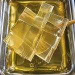 Transparent golden jelly cube lollipop arranged in a woven lattice pattern on a wooden stick over a tray of syrup.