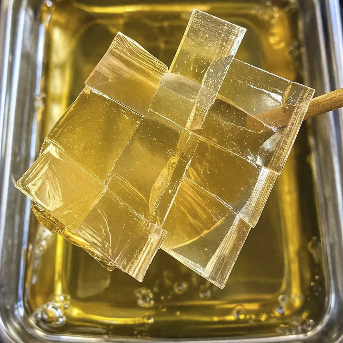 Transparent golden jelly cube lollipop arranged in a woven lattice pattern on a wooden stick over a tray of syrup.