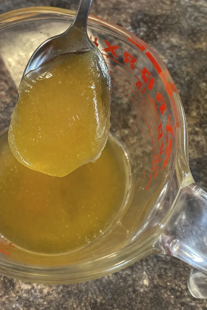 A spoon holding thick golden homemade gelatin or concentrated cooking liquid above a glass measuring cup in a kitchen setting.