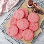 Overhead view of bright pink sugar cookies on parchment paper over a cooling rack with measuring spoons and vanilla extract.