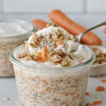 Creamy carrot cake overnight oats in a glass jar topped with yogurt and walnuts with a spoon lifting a bite