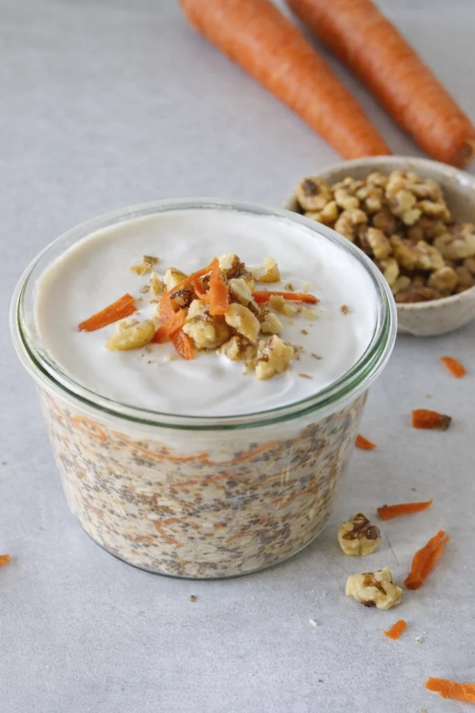 Carrot cake overnight oats in a glass jar topped with yogurt, walnuts, and shredded carrots with fresh ingredients in a clean kitchen setting