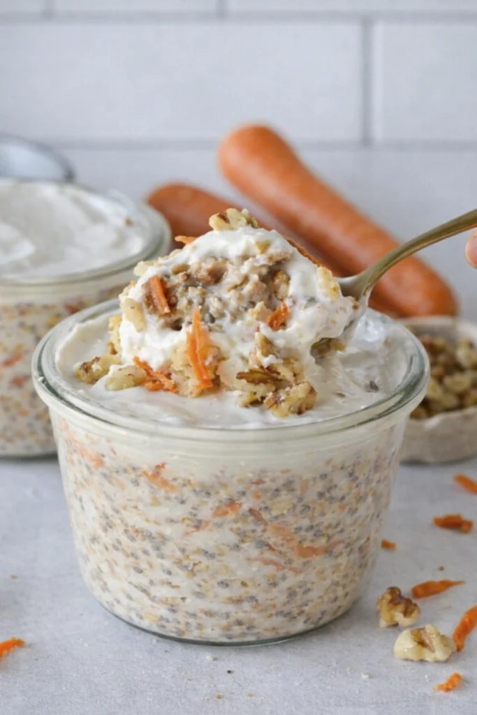 Close-up of carrot cake overnight oats in a glass jar with yogurt and walnuts, with a spoon lifting a creamy bite and fresh carrots in the background