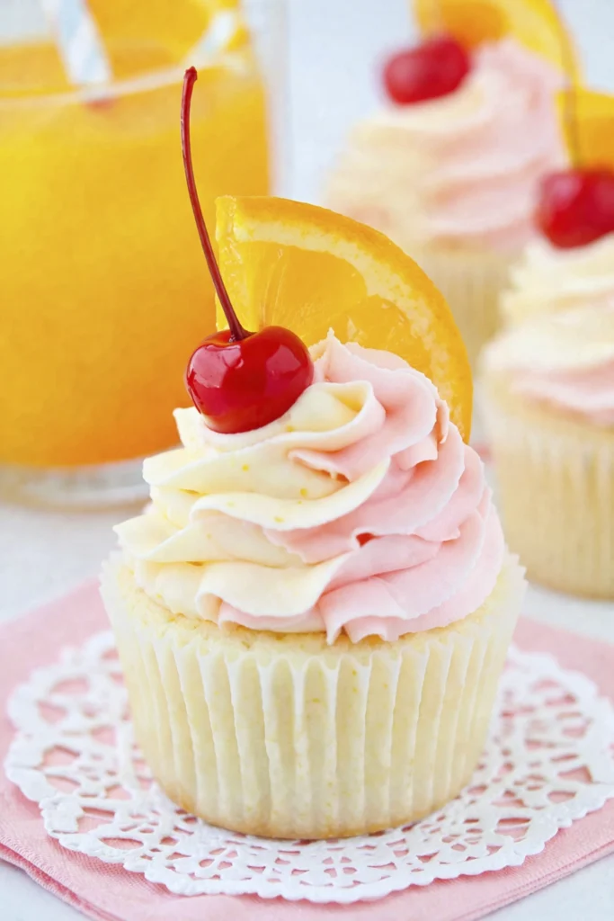 Close-up of a vanilla cupcake with pink buttercream frosting topped with a cherry and orange slice.