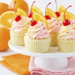 Orange creamsicle cupcakes with pink buttercream frosting, maraschino cherries, and fresh orange slices on a white cake stand.