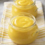 Two small glass jars filled with smooth and creamy lemon curd placed on a yellow striped cloth in soft natural light