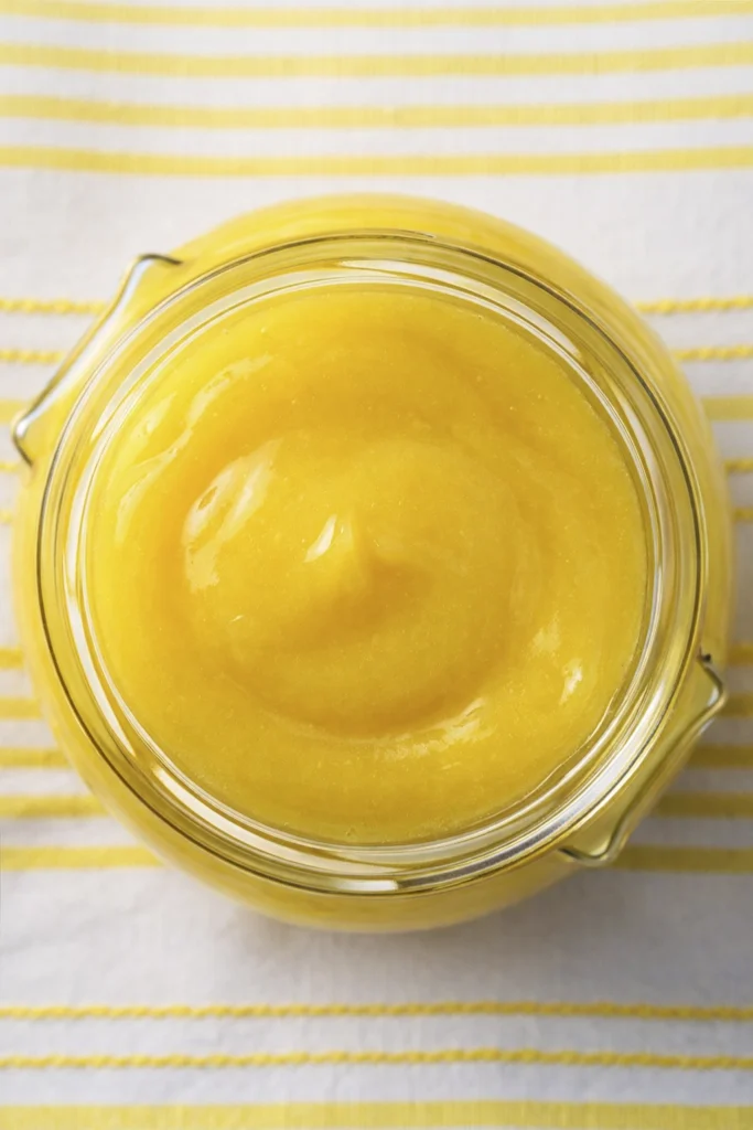 Top view of creamy lemon curd in a glass jar on a white cloth with yellow stripes.