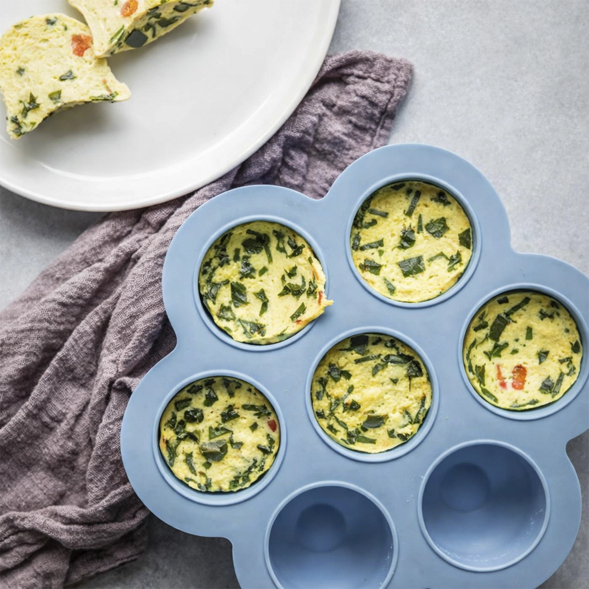 Baked egg bites in a blue silicone mold with herbs and vegetables, served with sliced egg muffins on a white plate