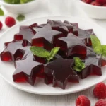 Glossy red star-shaped jelly dessert served on a white plate with fresh mint leaves and raspberries.