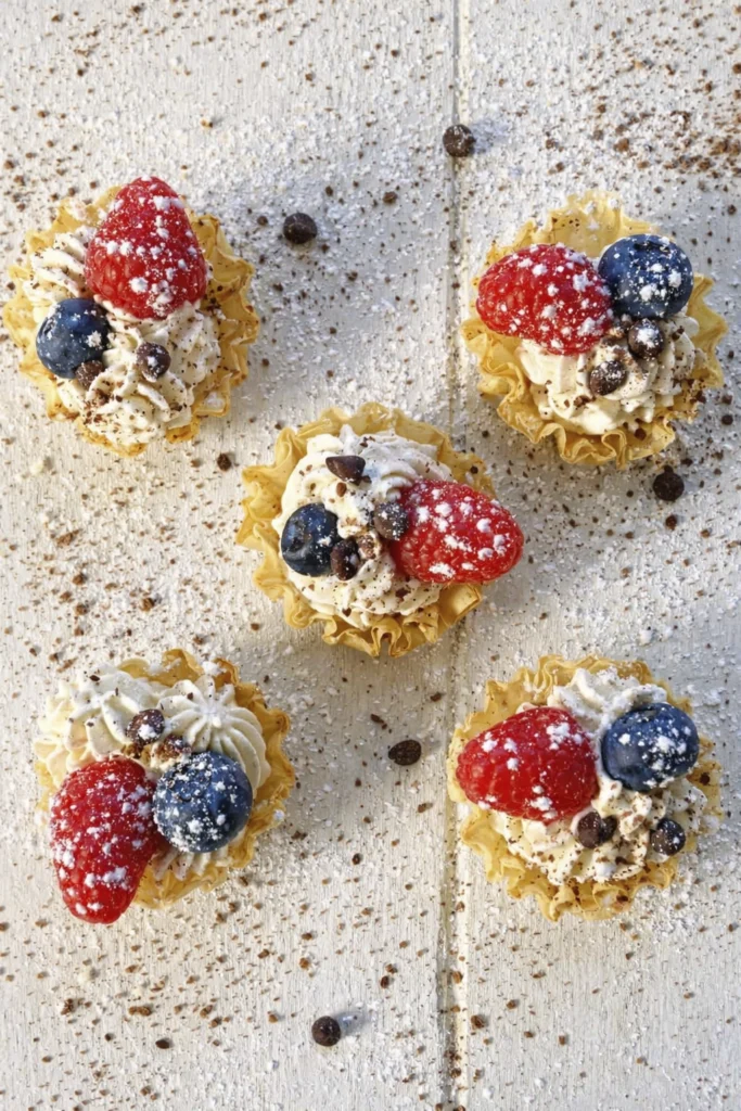 Top view of mini phyllo cups with whipped cream, raspberries, blueberries, and chocolate on a white wooden table