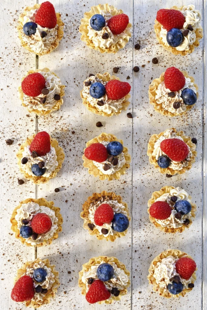 Mini phyllo pastry cups with whipped cream, berries, powdered sugar, and chocolate on rustic white table
